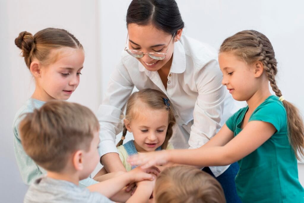teacher putting her hands together with her students game Çocuk Bakım Elemanı Sertifikası Nasıl Alınır? -