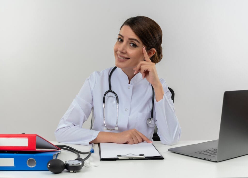 smiling young female doctor wearing medical robe stethoscope sitting desk with medical tools laptop touching head with finger isolated white wall Tıbbi Sekreterlik Sertifikası Nasıl Alınır?